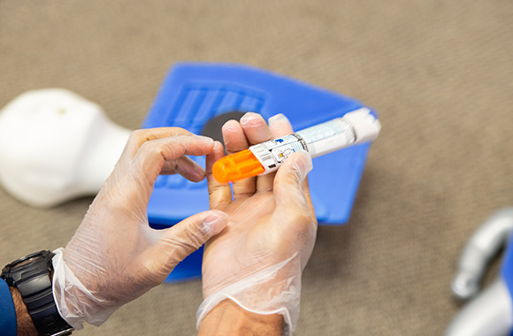 person wearing gloves holding an epinephrine injector in a training setting demonstrating emergency response techniques for allergic reactions and anaphylaxis management