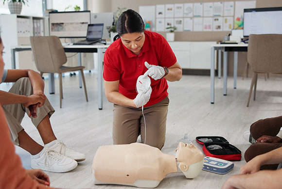 a woman in a red shirt demonstrating CPR techniques on a manikin in an educational setting surrounded by learners engaged in hands on training showing important life saving skills