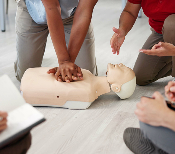 hands performing CPR on a training mannequin with observers nearby in a first aid demonstration focused on lifesaving techniques and practice sessions