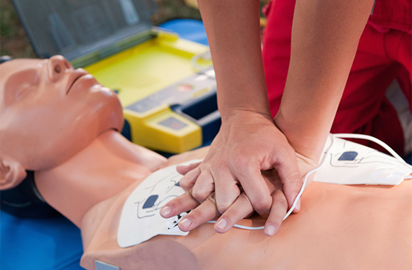 hands performing CPR on a training dummy with an automated external defibrillator visible in the background demonstrating lifesaving techniques for emergency response 2 steps to save a life